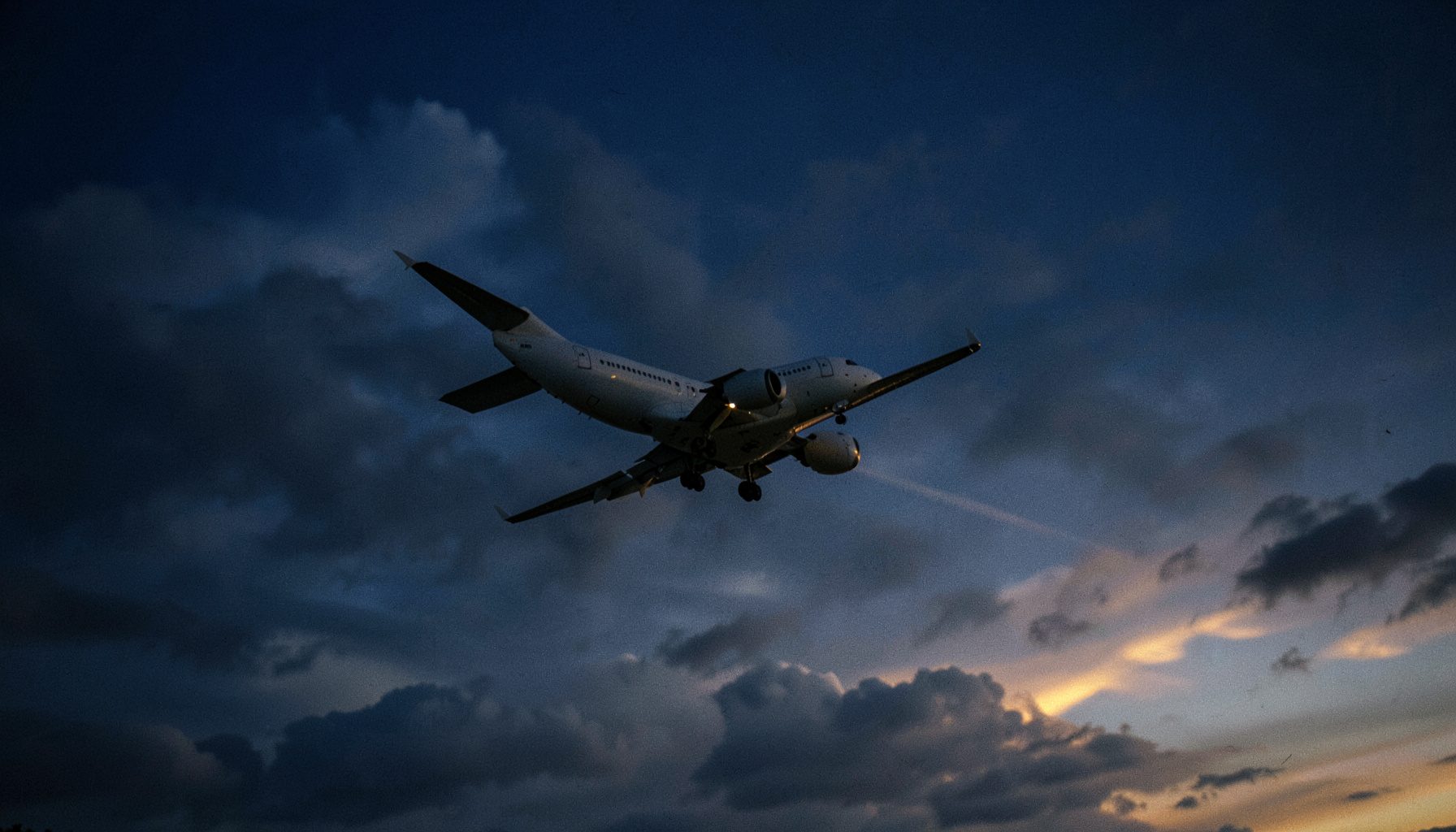 Wide-angle photograph of a commercial jet banking over a deep navy sky at dusk, steel-gray clouds behind it and warm horizon light at the edges, conveying motion and ascent in cool muted tones